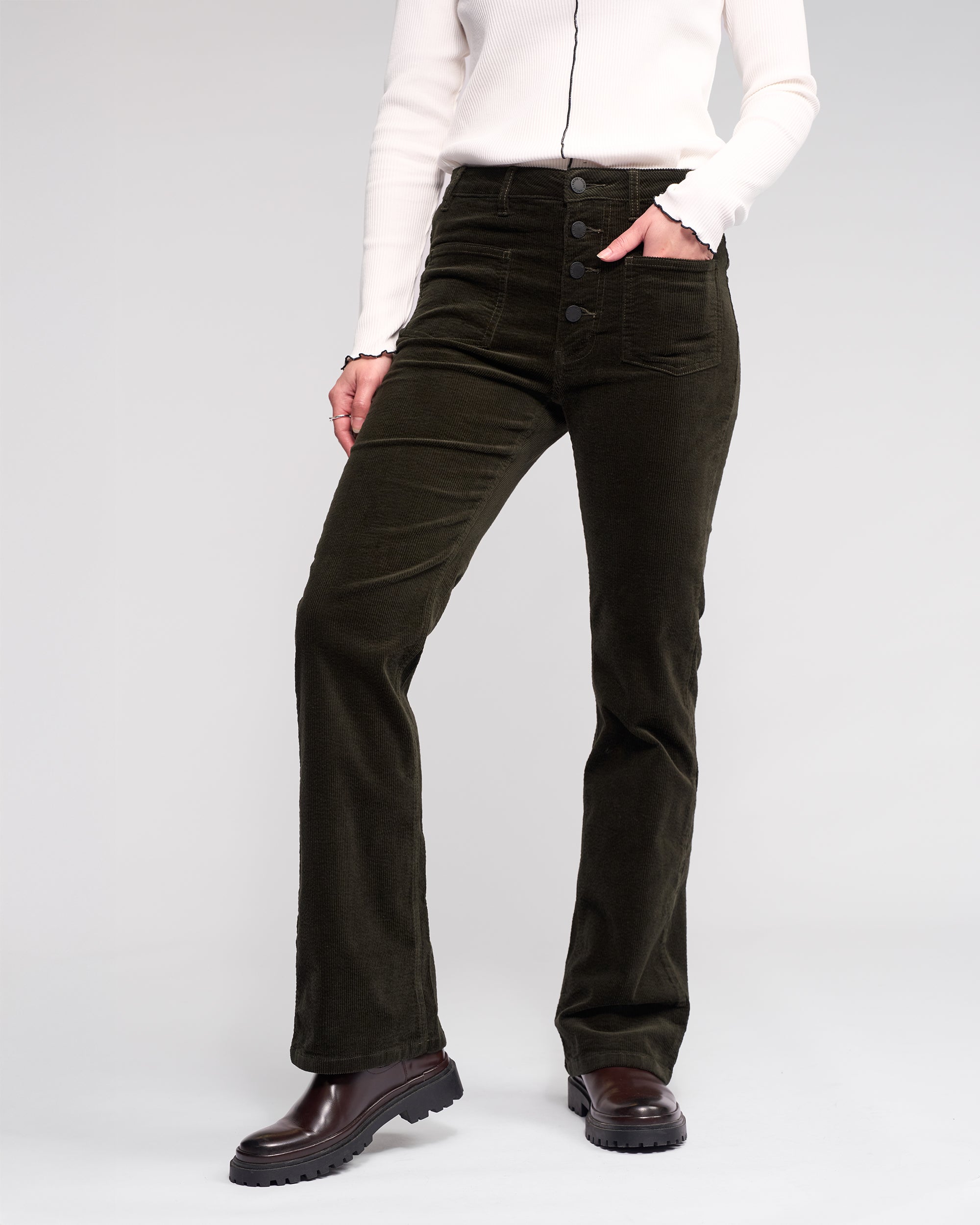 Wearing the Knewe Label Potter Cord - Hunter bootcut stretch wide-wale corduroy pants with a white long-sleeve top and dark brown chunky boots, a person stands against a plain light gray background.