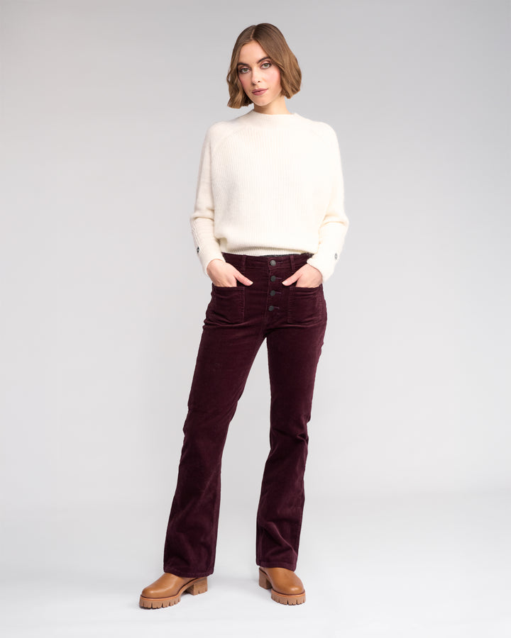A woman poses against a plain light background, wearing Knewe Label’s Potter Cord - Raisin bootcut corduroy pants with a cream sweater and brown platform shoes, her hands in her pockets as she gazes slightly to the side.