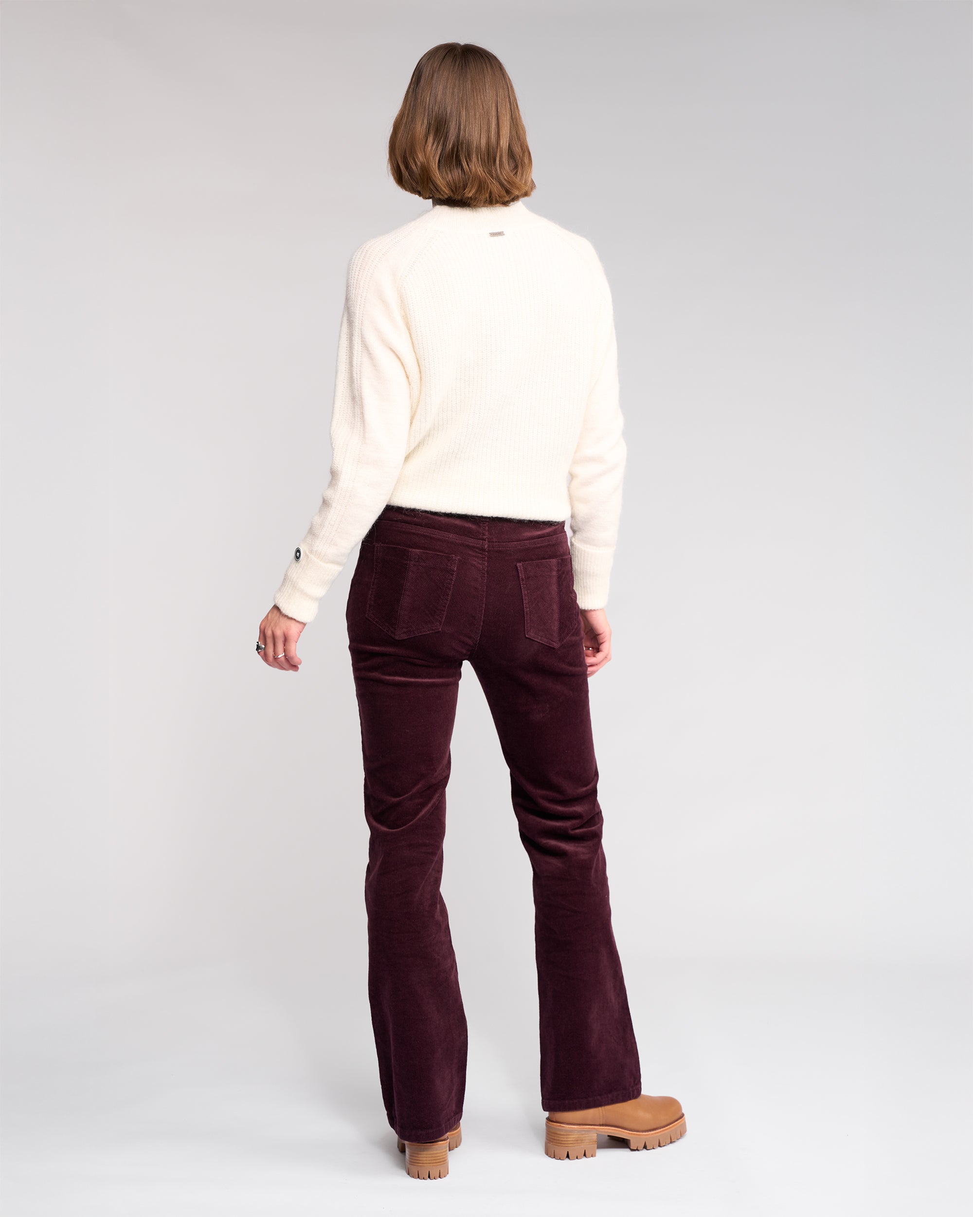 A person with shoulder-length hair stands facing away, wearing a cream sweater, tan platform shoes, and the Knewe Label Potter Cord - Raisin bootcut corduroy pants in stretch wide-wale cord, against a plain light background.