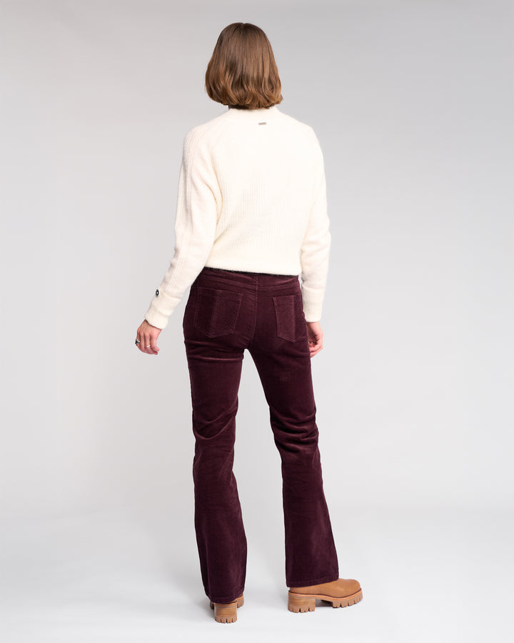 A person with shoulder-length hair stands facing away, wearing a cream sweater, tan platform shoes, and the Knewe Label Potter Cord - Raisin bootcut corduroy pants in stretch wide-wale cord, against a plain light background.