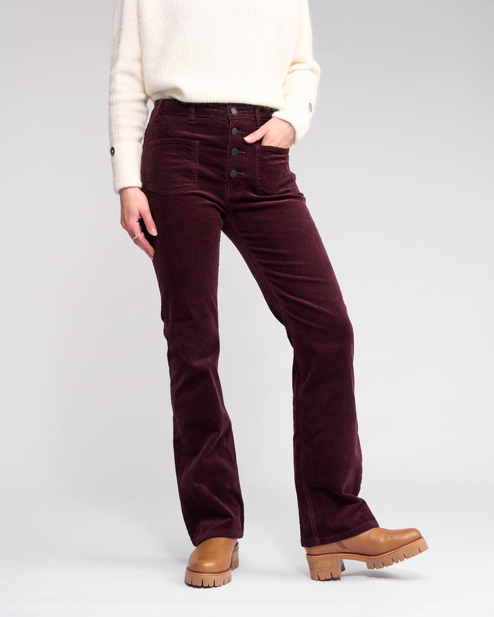 A person wears Knewe Label's Potter Cord - Raisin high-waisted bootcut pants with buttons, a white sweater, and tan platform shoes, posing with one hand in their pocket against a plain backdrop.