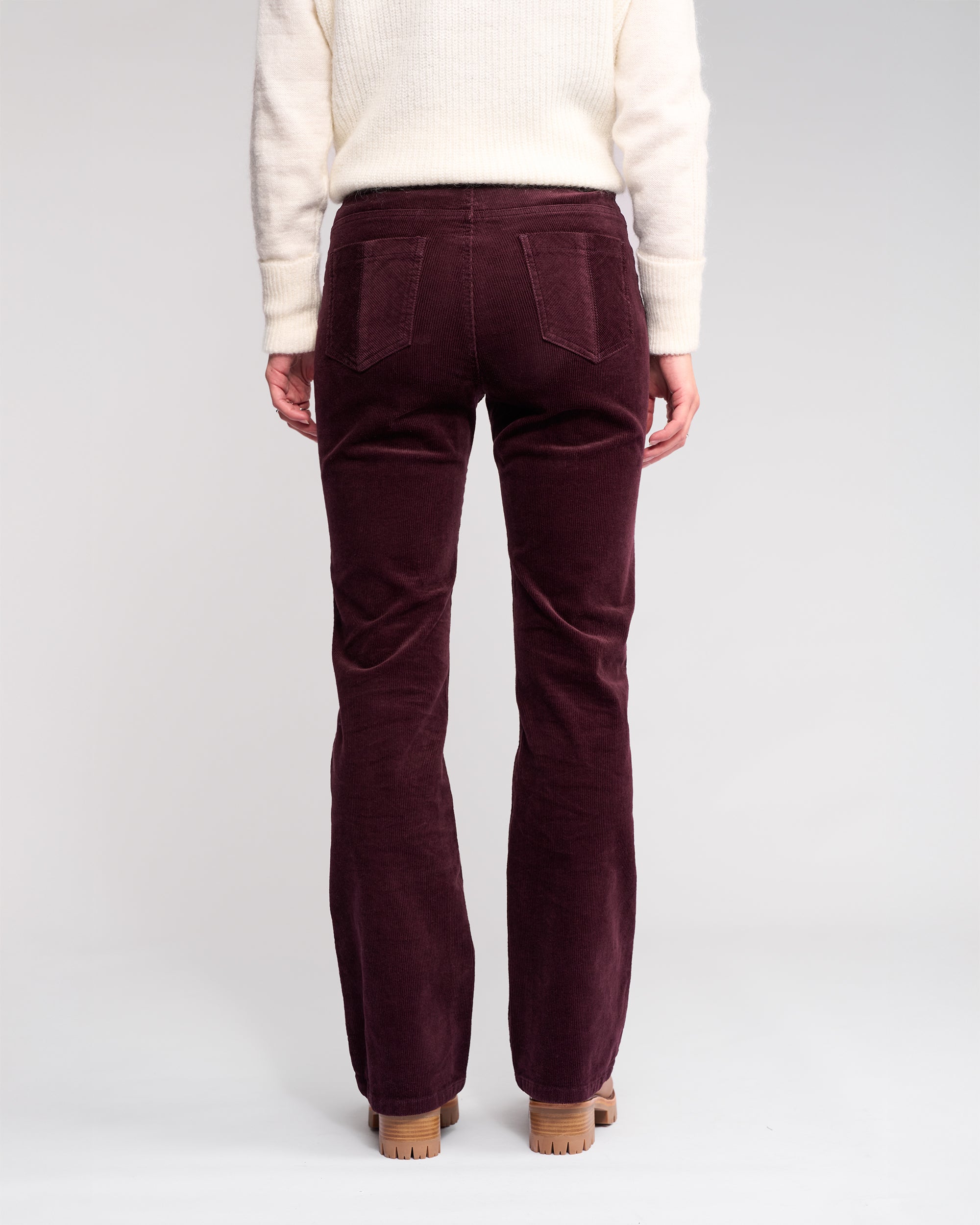Only the lower half of a person is shown from the back, wearing Knewe Label's Potter Cord - Raisin bootcut pants, made from stretch wide-wale cord, paired with a cream sweater on a light studio background.