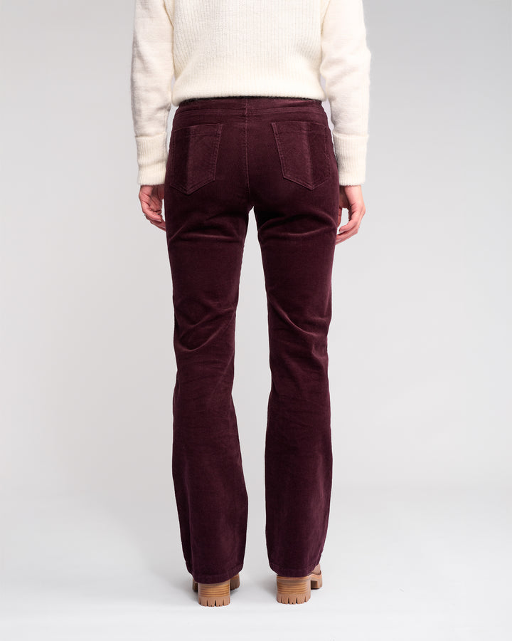 Only the lower half of a person is shown from the back, wearing Knewe Label's Potter Cord - Raisin bootcut pants, made from stretch wide-wale cord, paired with a cream sweater on a light studio background.