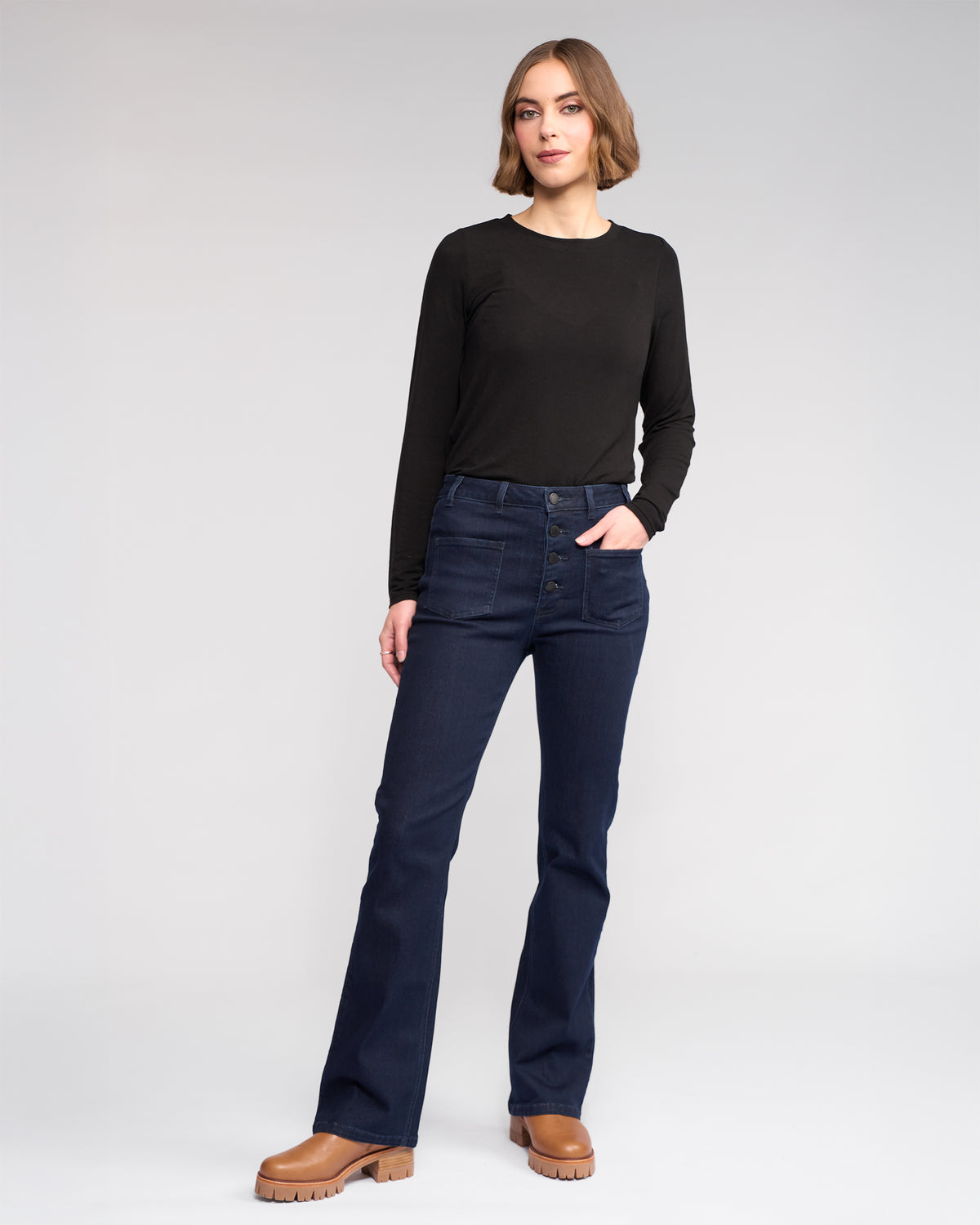 A person stands against a light background wearing Knewe Label's Potter Jean in Ink Denim, a high-waisted stretch bootcut style with buttons, paired with a black long-sleeve shirt and brown shoes, one hand in their pocket and looking relaxed.