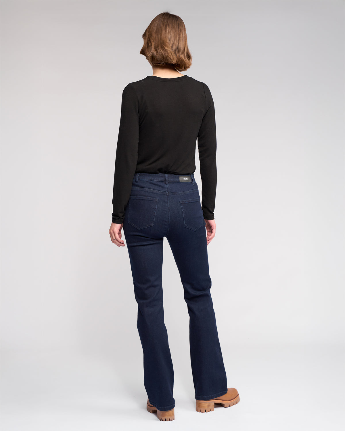A person with short brown hair, facing away from the camera on a plain light background, wears a long-sleeved black shirt and Knewe Label's Potter Jean in Ink Denim.