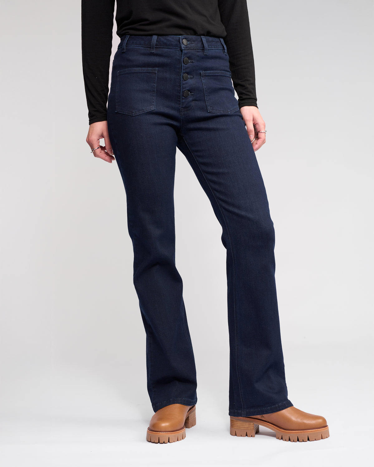 A person wears the Knewe Label Potter Jean in Ink Denim—high-waisted, straight-leg jeans with front buttons—paired with a black long-sleeve top and tan platform shoes, standing against a plain light background.