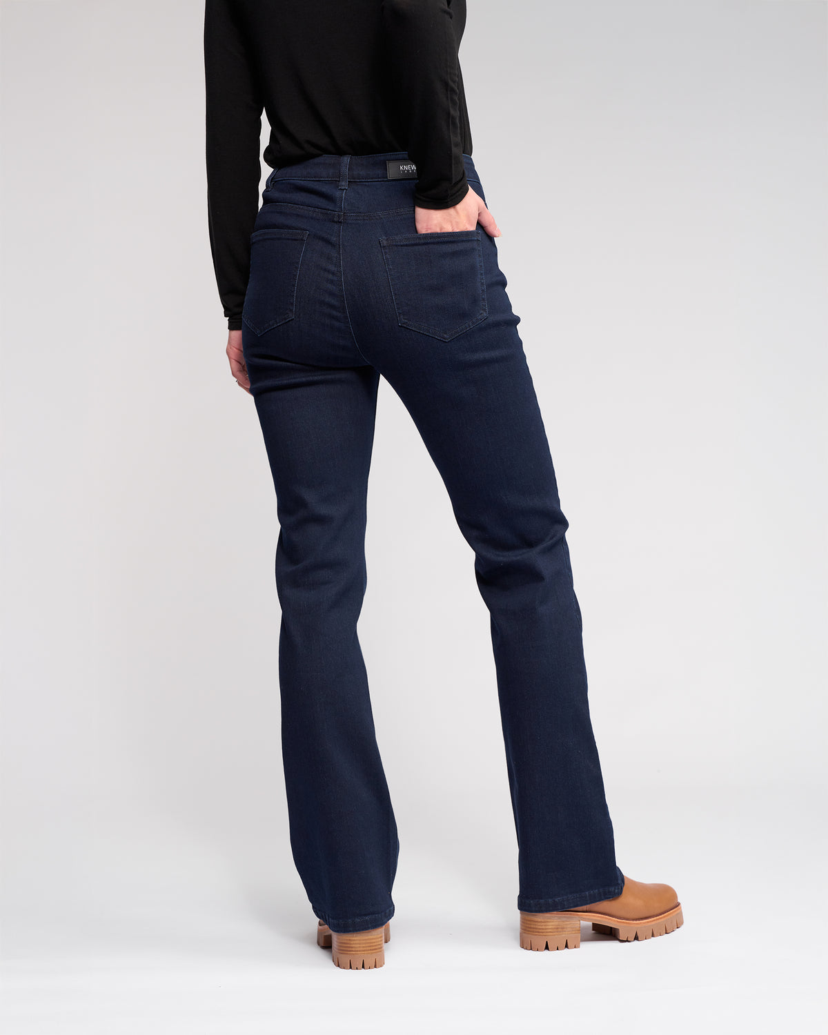 Wearing the Knewe Label Potter Jean in Ink Denim, a black long-sleeve top, and tan chunky-heeled boots, a person stands with their back to the camera against a plain light background.