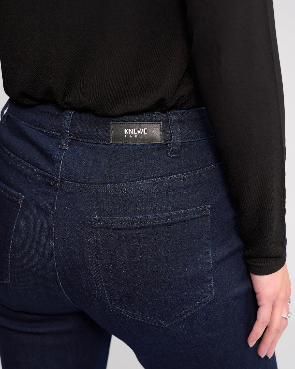 A person wears the Potter Jean - Ink Denim by Knewe Label, a dark blue bootcut stretch denim with a branded waistband label above the right back pocket. The person’s left hand hangs by their side.