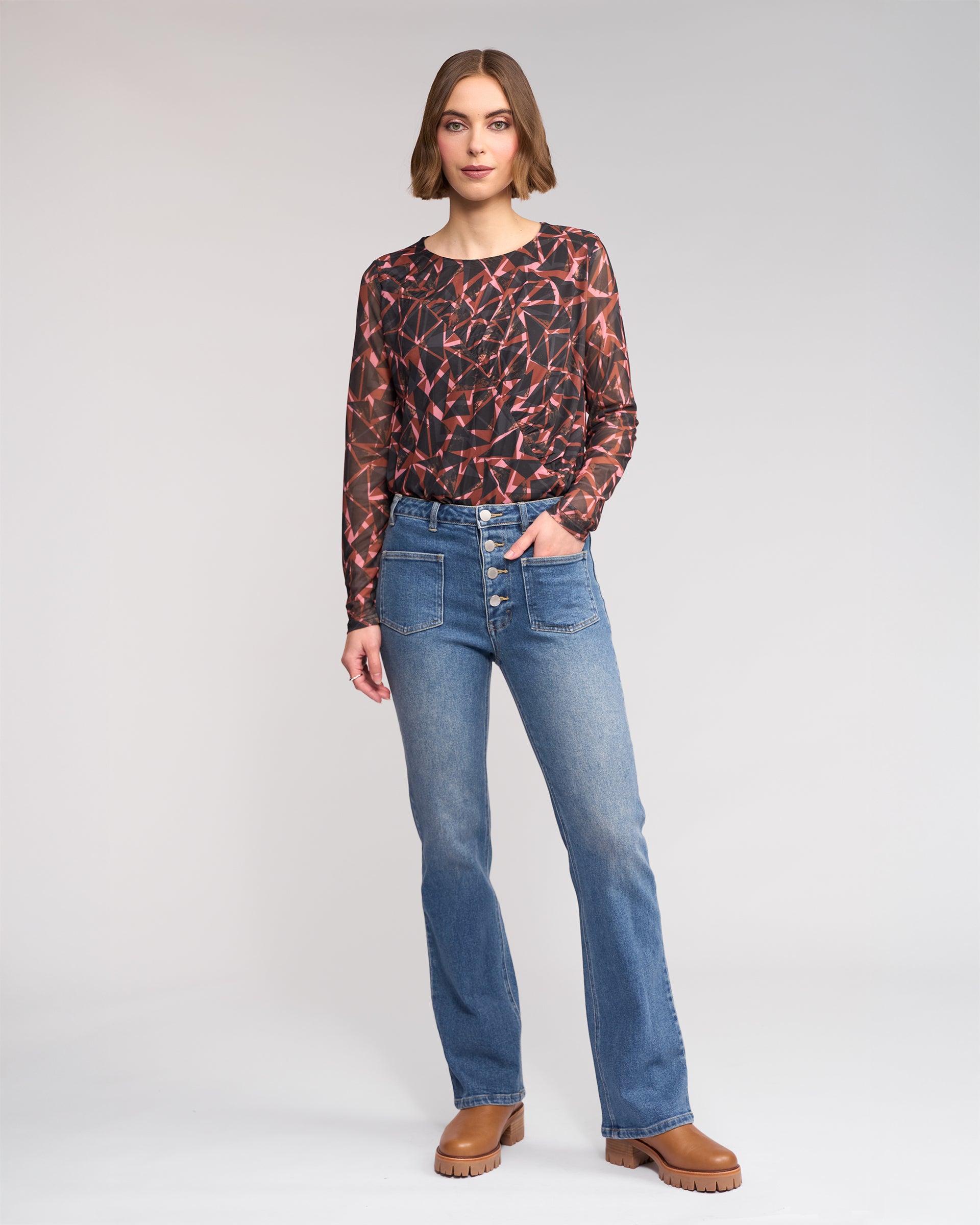 A woman stands against a plain background wearing the Knewe Label Potter Jean - Vintage Denim, styled with a patterned long-sleeve top and brown boots. One hand is in her pocket as she looks at the camera.