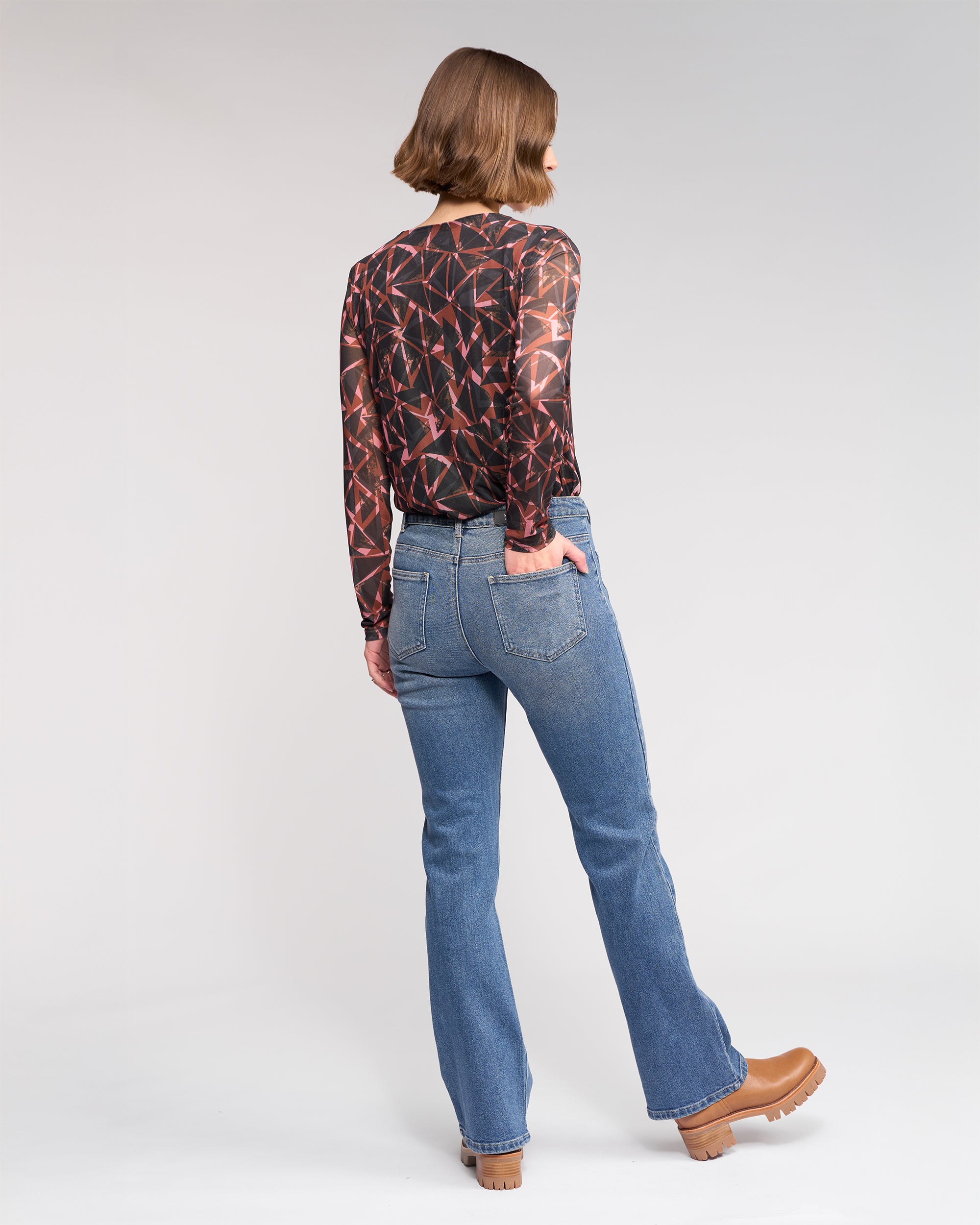 A person with short brown hair faces away, wearing a black and red geometric-patterned long-sleeve top, Knewe Label's Potter Jean in Vintage Denim, and tan platform shoes against a plain light background.