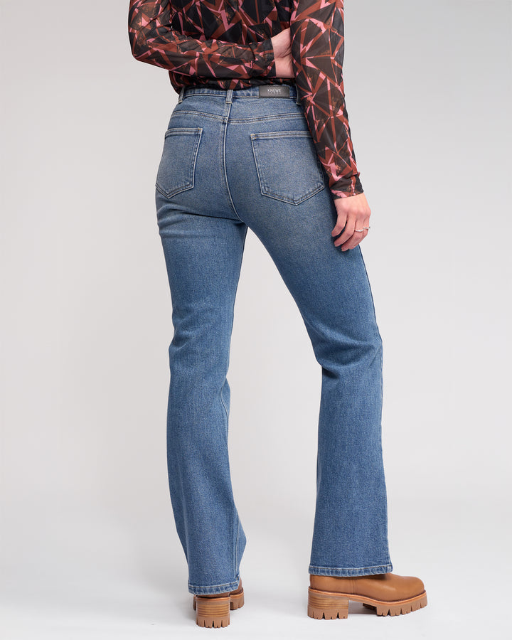 A person faces away, wearing Knewe Label's Potter Jean in vintage denim, a long-sleeve geometric patterned top, and tan chunky-heeled boots against a plain light background.