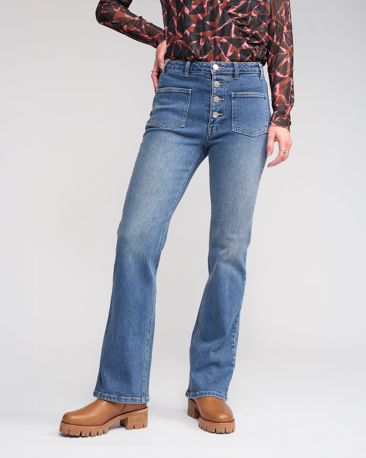 A person stands against a plain background wearing Knewe Label's Potter Jean - Vintage Denim, styled with a patterned long-sleeve top and brown platform shoes.