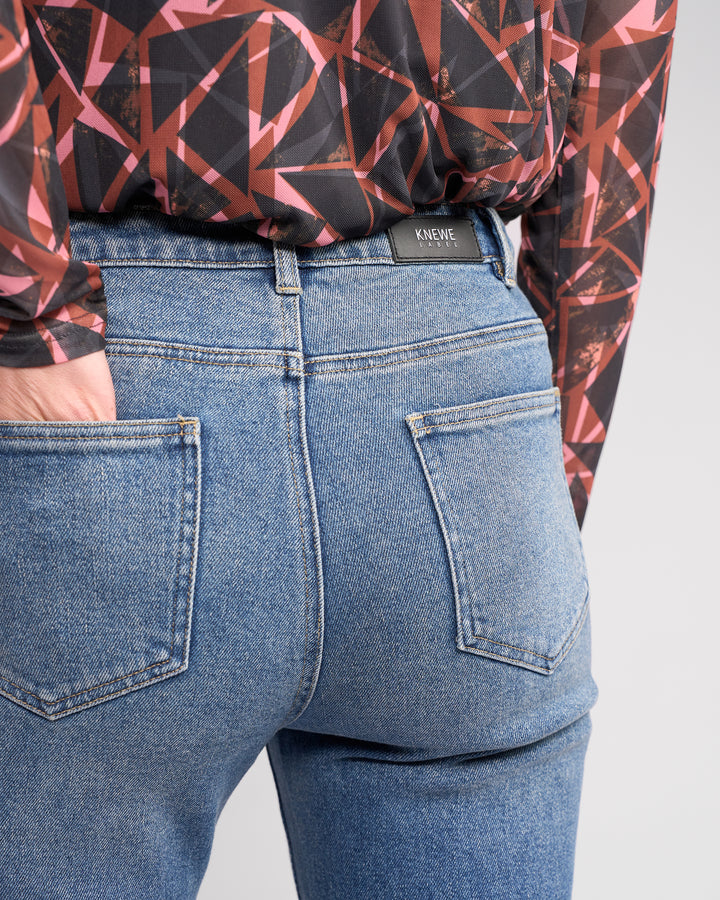 A person wears the Potter Jean - Vintage Denim by Knewe Label with a geometric-patterned shirt, standing with one hand in a back pocket. The bootcut jeans display the Knewe Label above the right pocket.