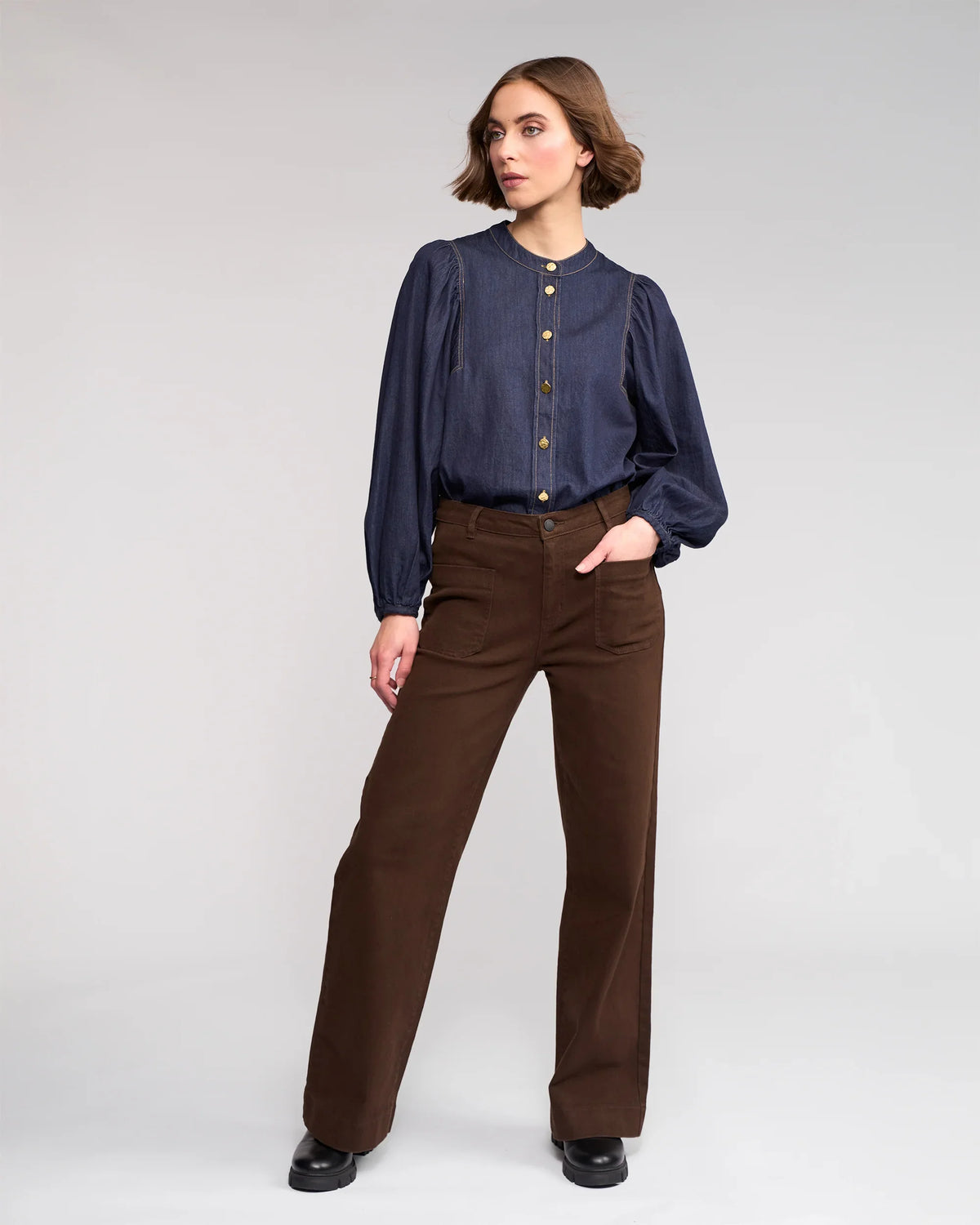 A woman with short hair stands against a plain background, wearing the Knewe Label Roxie Jean in chocolate, paired with a navy long-sleeve puff-sleeve blouse and black shoes. She looks off to the side with one hand in her pocket.