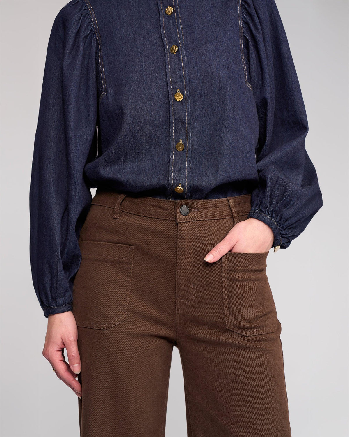 A person stands with one hand in the pocket, wearing high-waisted Knewe Label Roxie Jean in Chocolate and a long-sleeved dark blue button-up shirt. The head is not visible.