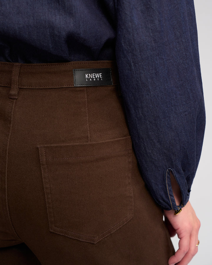 Someone wearing the Knewe Label Roxie Jean in chocolate, featuring patch pockets and a black brand tag, paired with a navy blue long-sleeve shirt.