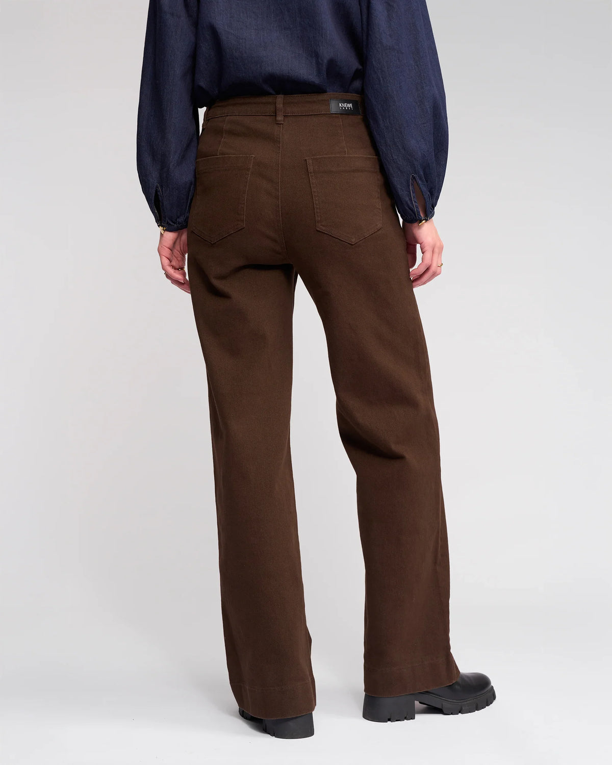 Someone wearing the Knewe Label Roxie Jean in chocolate, paired with a dark long-sleeve shirt and black shoes, is shown from the back against a plain light background.