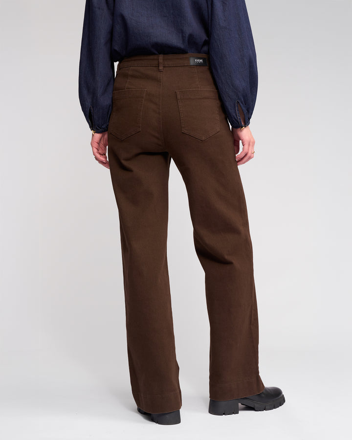 Someone wearing the Knewe Label Roxie Jean in chocolate, paired with a dark long-sleeve shirt and black shoes, is shown from the back against a plain light background.