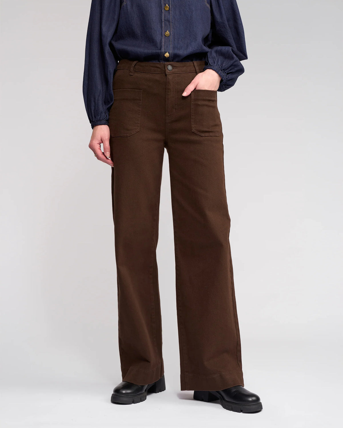 A person stands against a plain background wearing Knewe Label's Roxie Jean in Chocolate, featuring a high waist, wide legs, and patch pockets. The dark blue long-sleeve shirt is tucked in, with black shoes. The face is not visible.