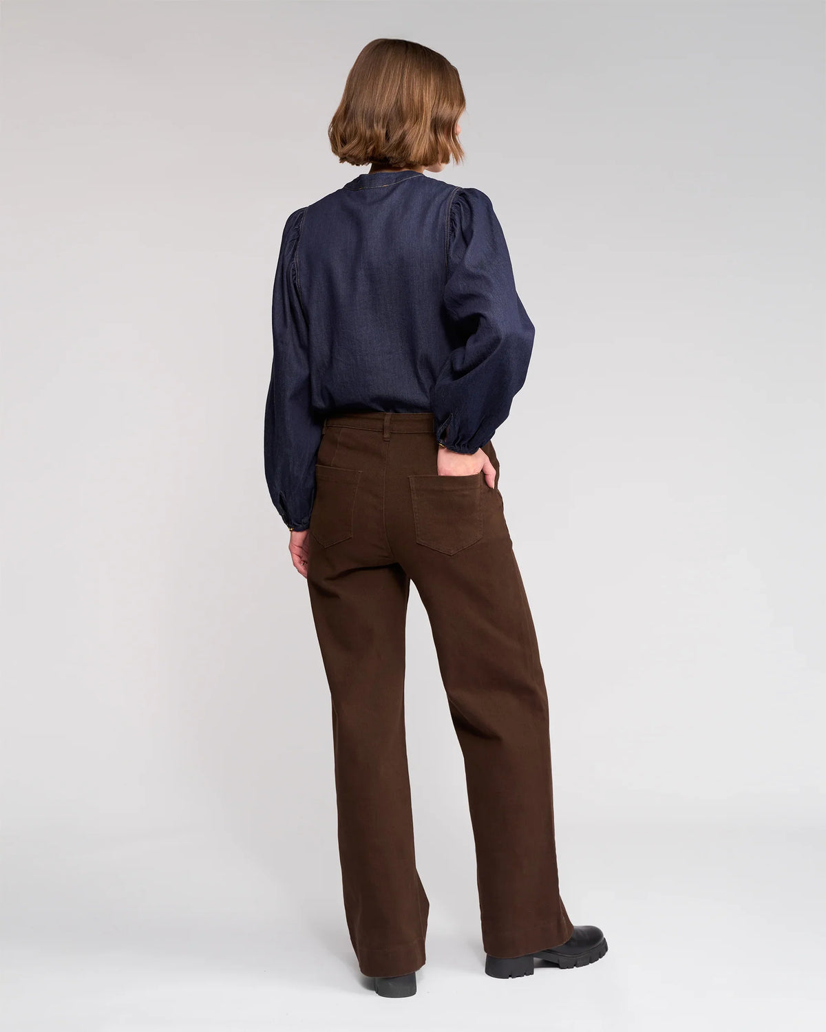 A person with short, light brown hair faces away, wearing a dark navy long-sleeve blouse and Knewe Label’s Roxie Jean - Chocolate, high-waisted wide-leg jeans with patch pockets, paired with black shoes against a plain light background.