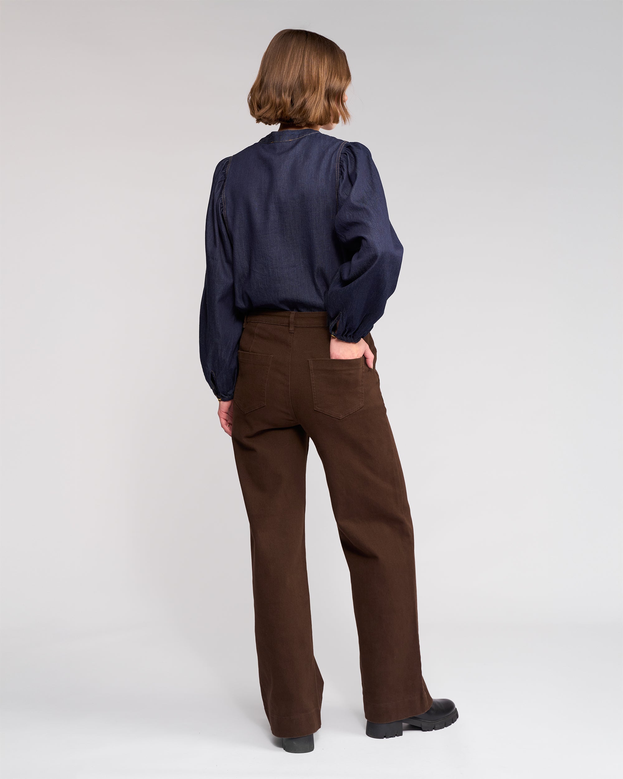 A person with short, light brown hair faces away, wearing a dark navy long-sleeve blouse and Knewe Label’s Roxie Jean - Chocolate, high-waisted wide-leg jeans with patch pockets, paired with black shoes against a plain light background.
