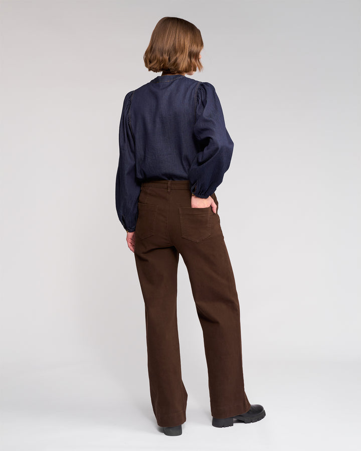 A person with short, light brown hair faces away, wearing a dark navy long-sleeve blouse and Knewe Label’s Roxie Jean - Chocolate, high-waisted wide-leg jeans with patch pockets, paired with black shoes against a plain light background.