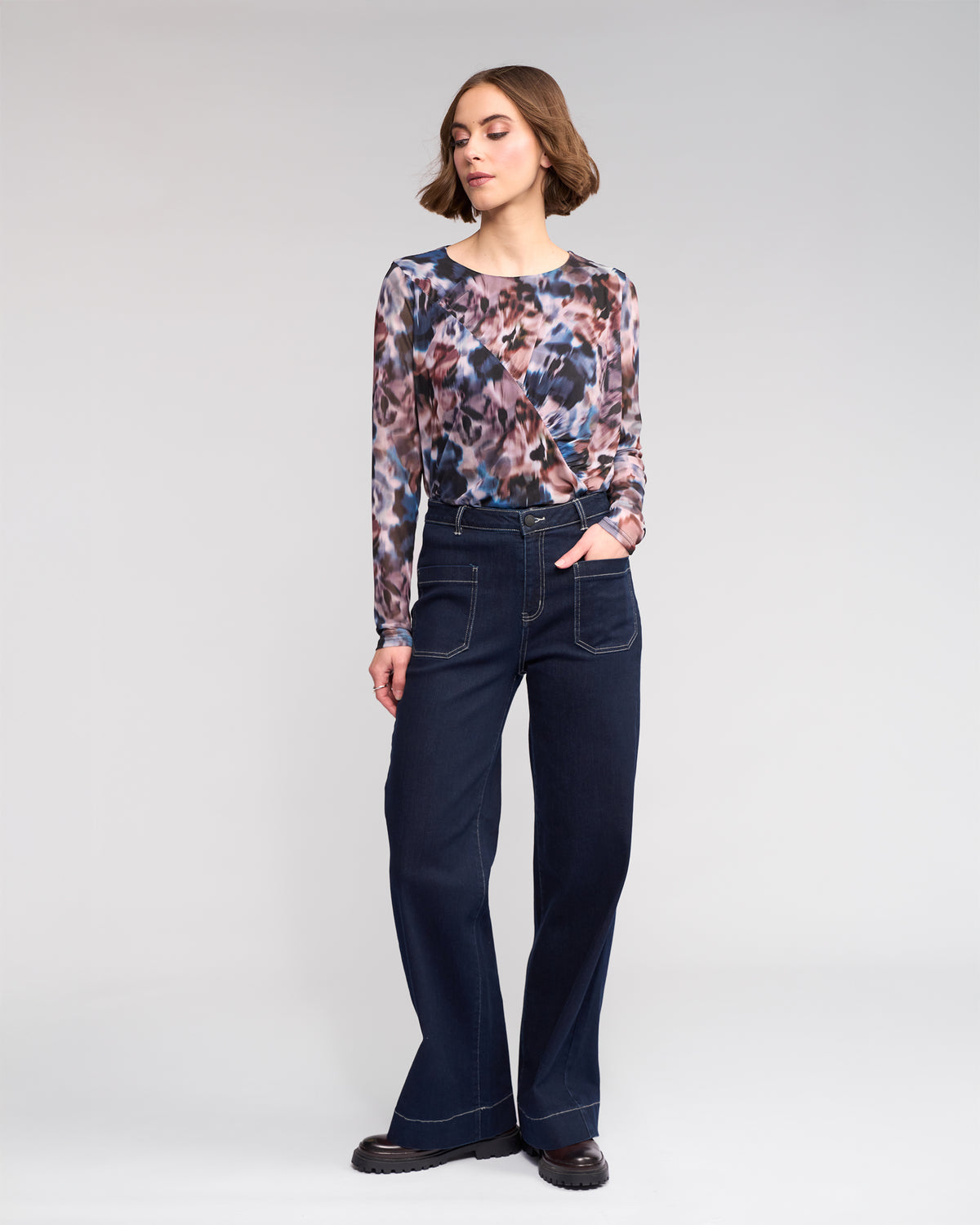 A woman stands against a plain background, wearing a long-sleeve, abstract-patterned top, high-waisted wide-leg dark jeans with front pockets, and black shoes. She has short brown hair and one hand in her pocket.