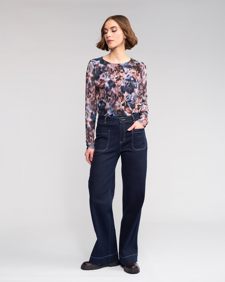 A woman stands against a plain background, wearing a long-sleeve, abstract-patterned top, high-waisted wide-leg dark jeans with front pockets, and black shoes. She has short brown hair and one hand in her pocket.