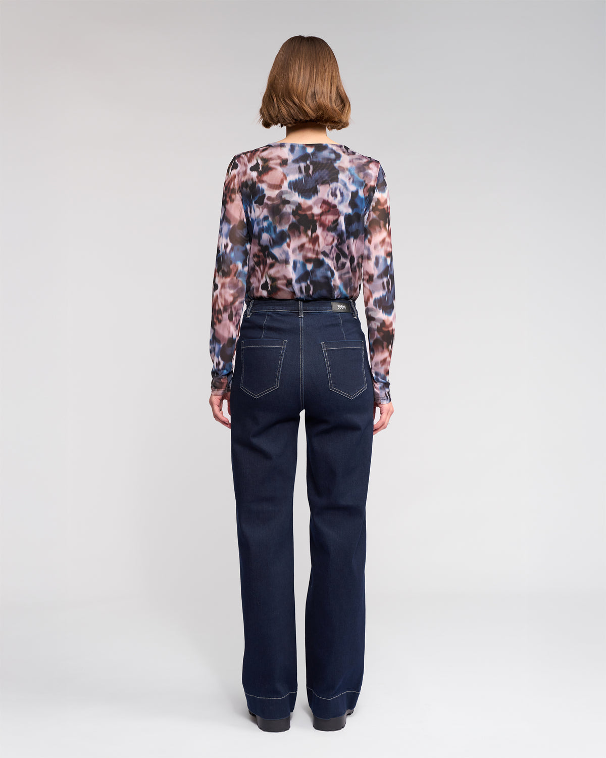 A person with short brown hair, seen from the back, wears a long-sleeve multicolored patterned blouse and high-waisted dark blue jeans with white stitching, standing against a plain light background.