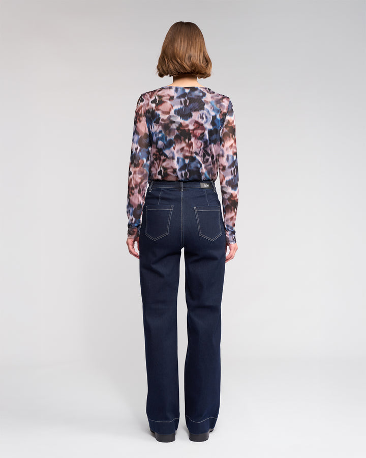 A person with short brown hair, seen from the back, wears a long-sleeve multicolored patterned blouse and high-waisted dark blue jeans with white stitching, standing against a plain light background.