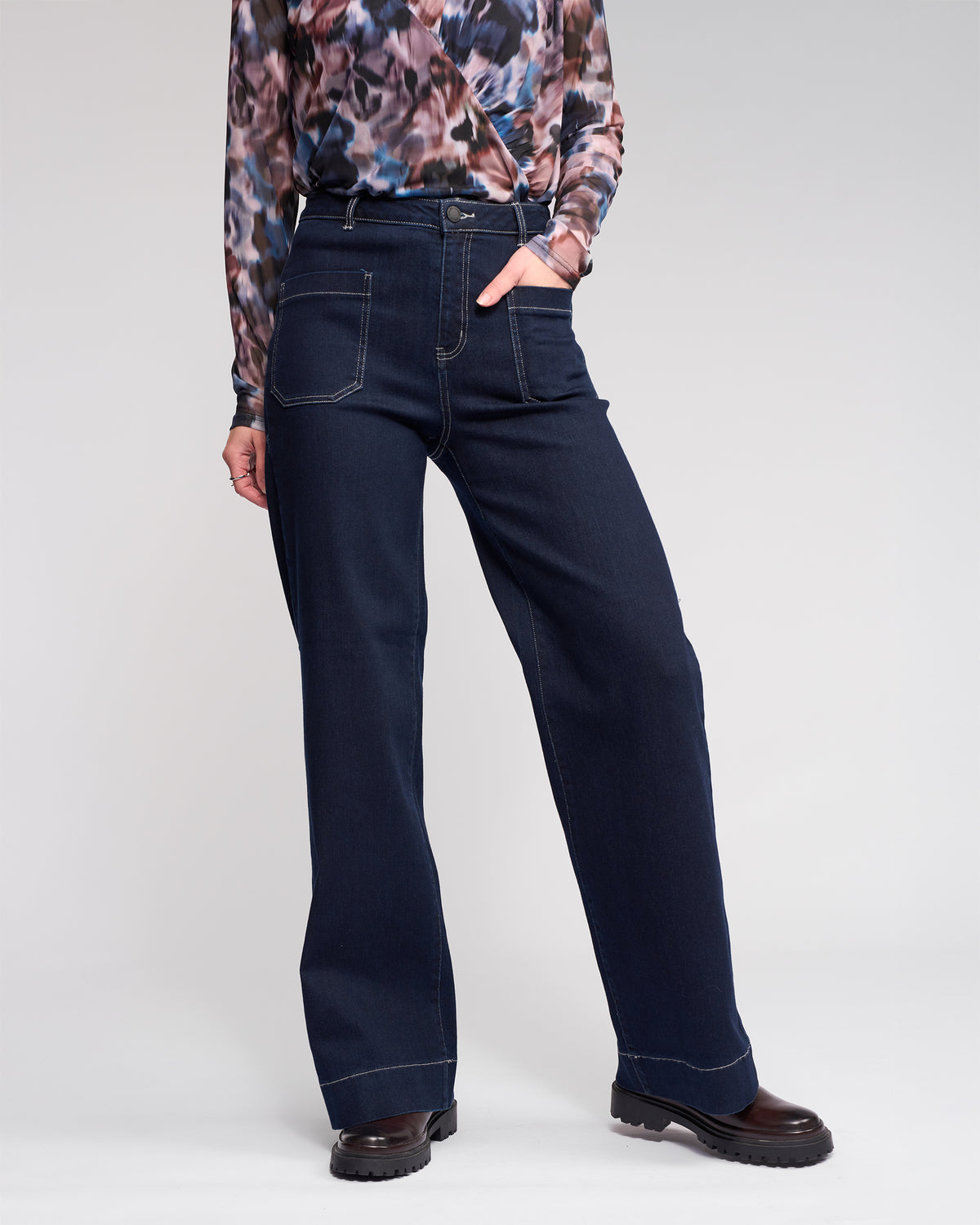 A person wears high-waisted, wide-leg dark blue jeans with prominent front pockets, a patterned long-sleeve top, and black chunky shoes, standing with one hand in a pocket against a light background.