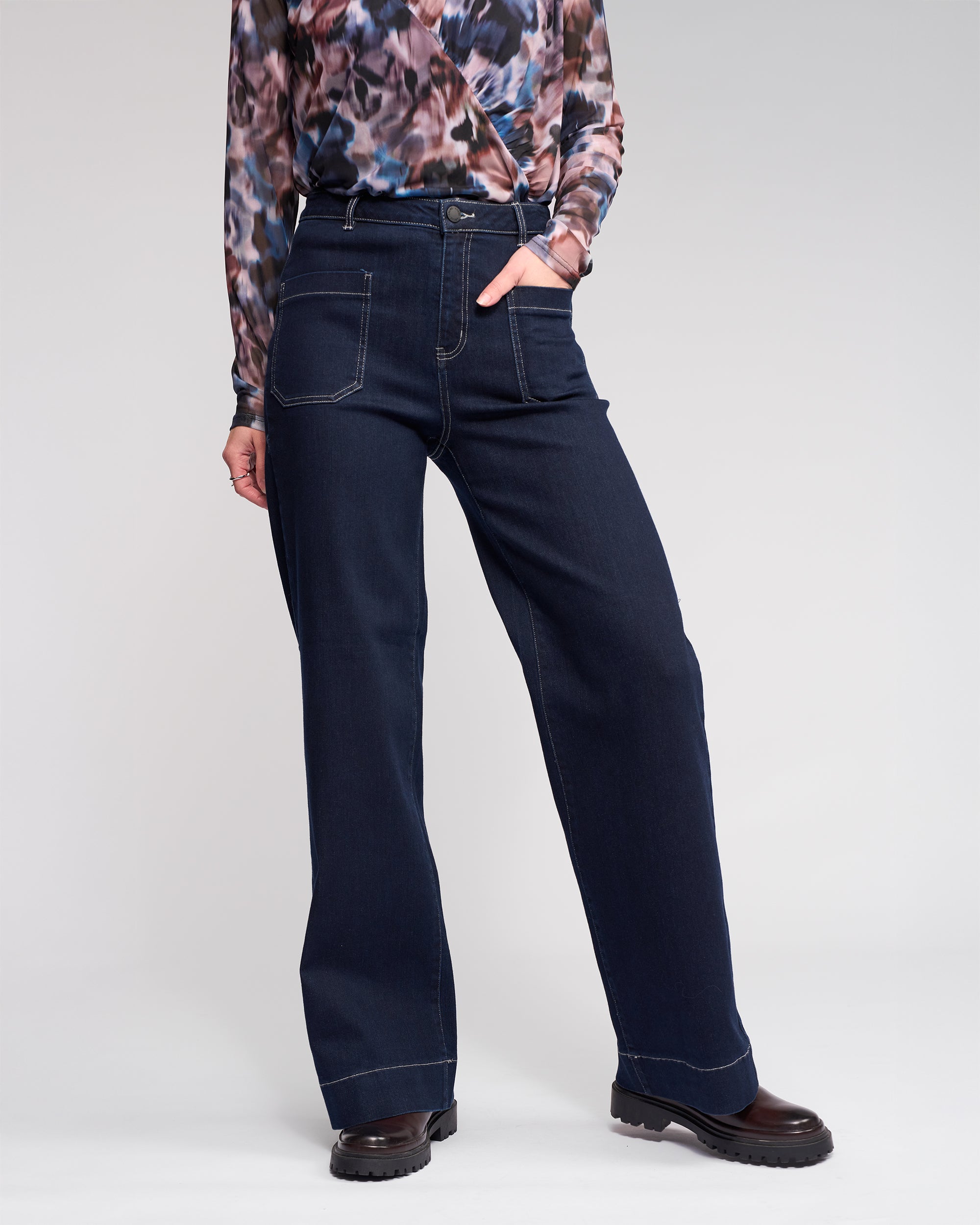 A person wears high-waisted, wide-leg dark blue jeans with prominent front pockets, a patterned long-sleeve top, and black chunky shoes, standing with one hand in a pocket against a light background.