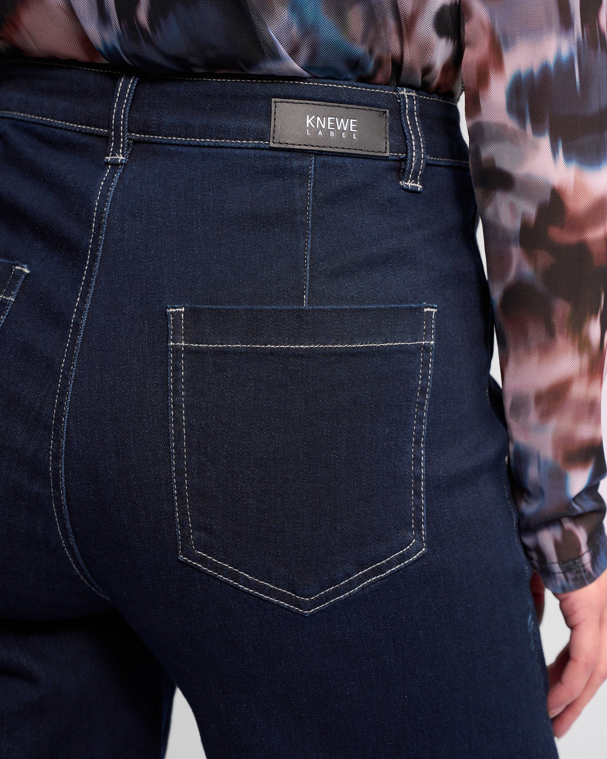 Close-up of the back of a person wearing dark blue jeans with white stitching and a black KNEWE label above the pocket. The person is also wearing a patterned long-sleeve shirt.
