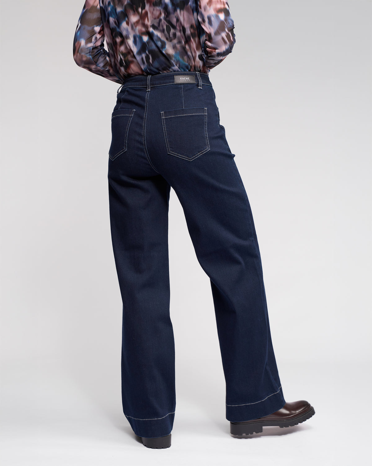 A person stands facing away, wearing dark blue wide-leg jeans, a patterned long-sleeve top, and black shoes against a plain light background.