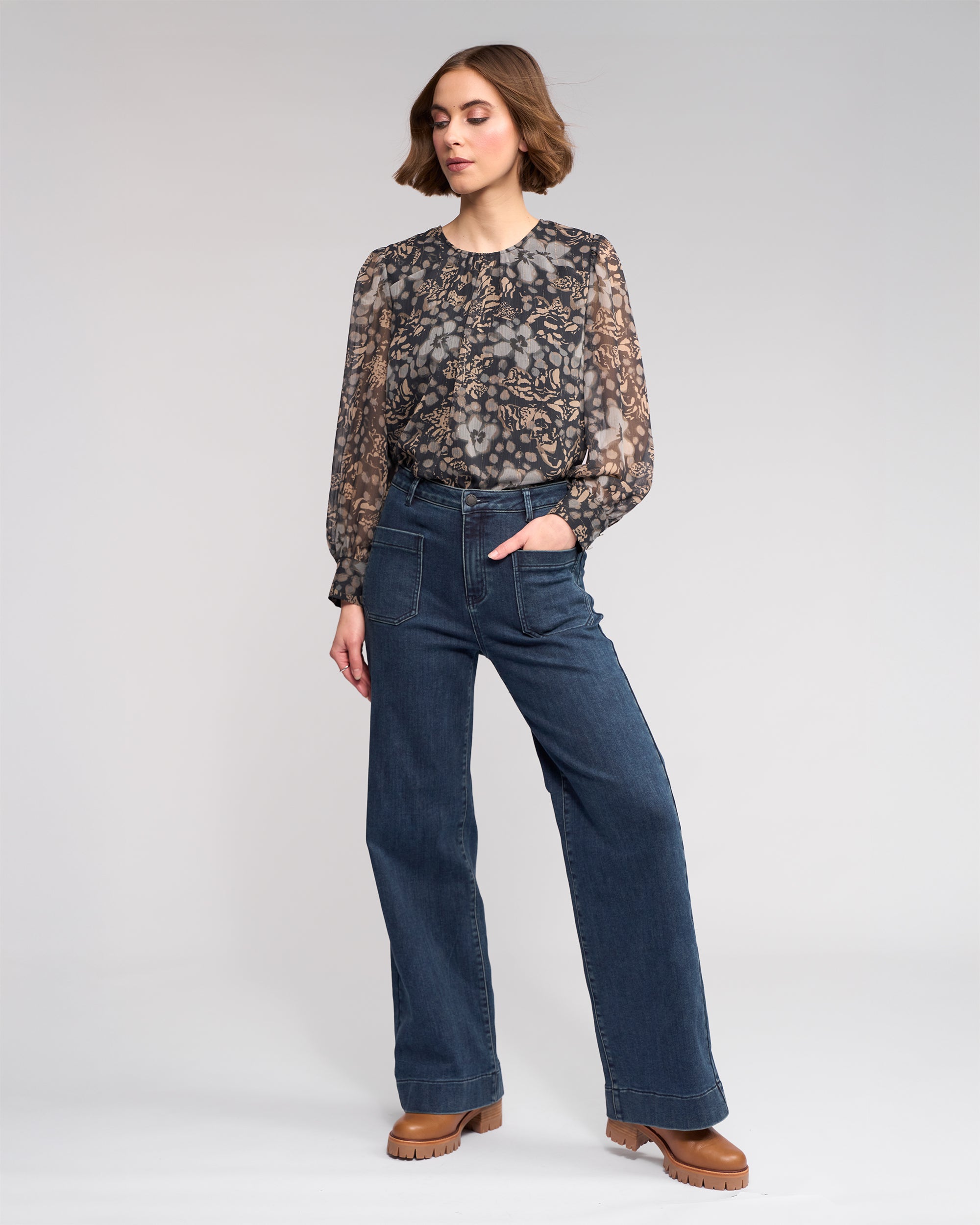 A woman poses with one hand in her pocket, wearing a sheer patterned blouse, Knewe Label Roxie Jean in Slate Denim wide-leg style, and brown platform shoes against a plain light background.
