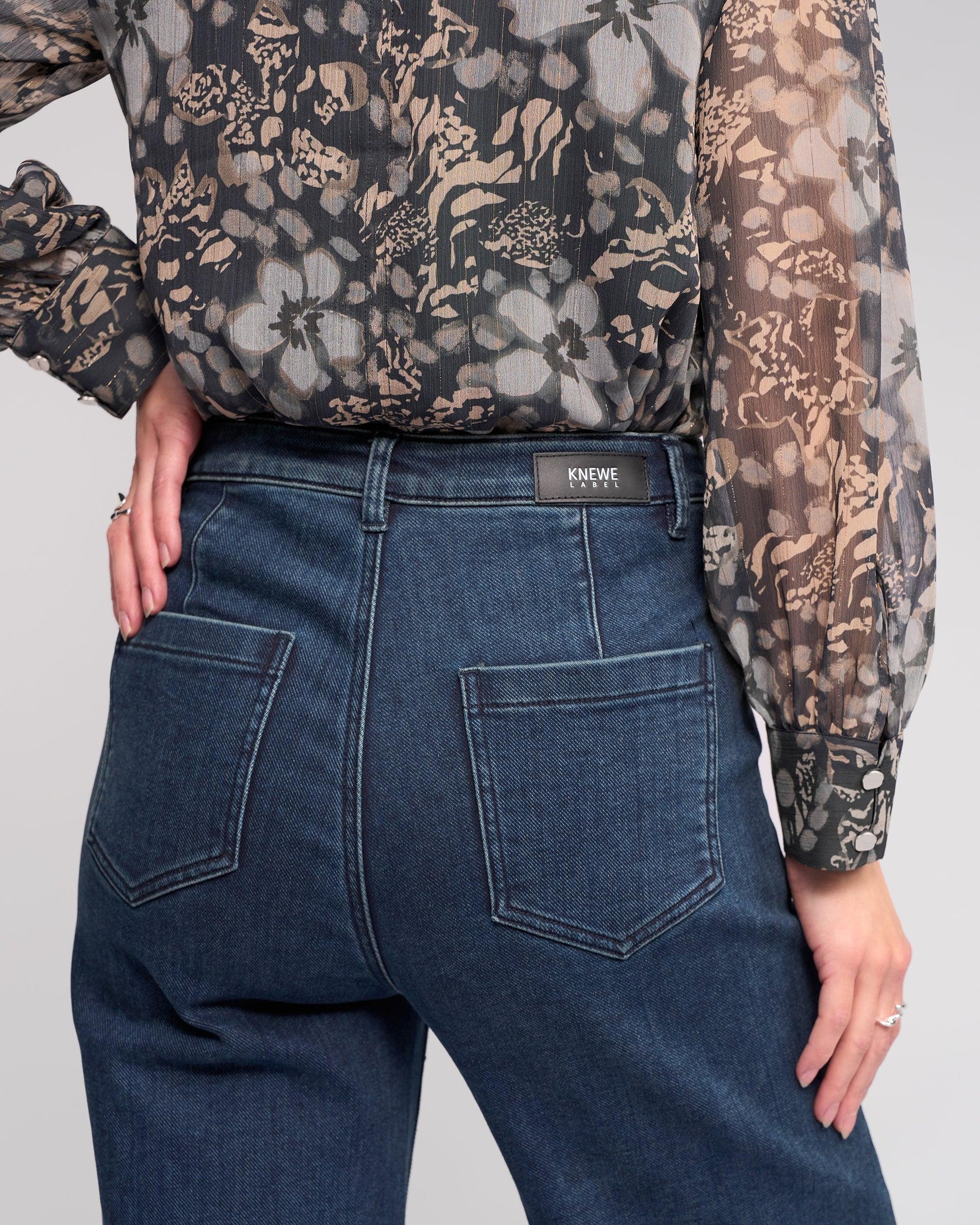 A person wears a sheer, patterned blouse and Knewe Label’s Roxie Jean in Slate Denim—high-waisted, wide-leg jeans with visible patch pockets and a KNEW label on the waistband—shown from behind, one hand on their hip.