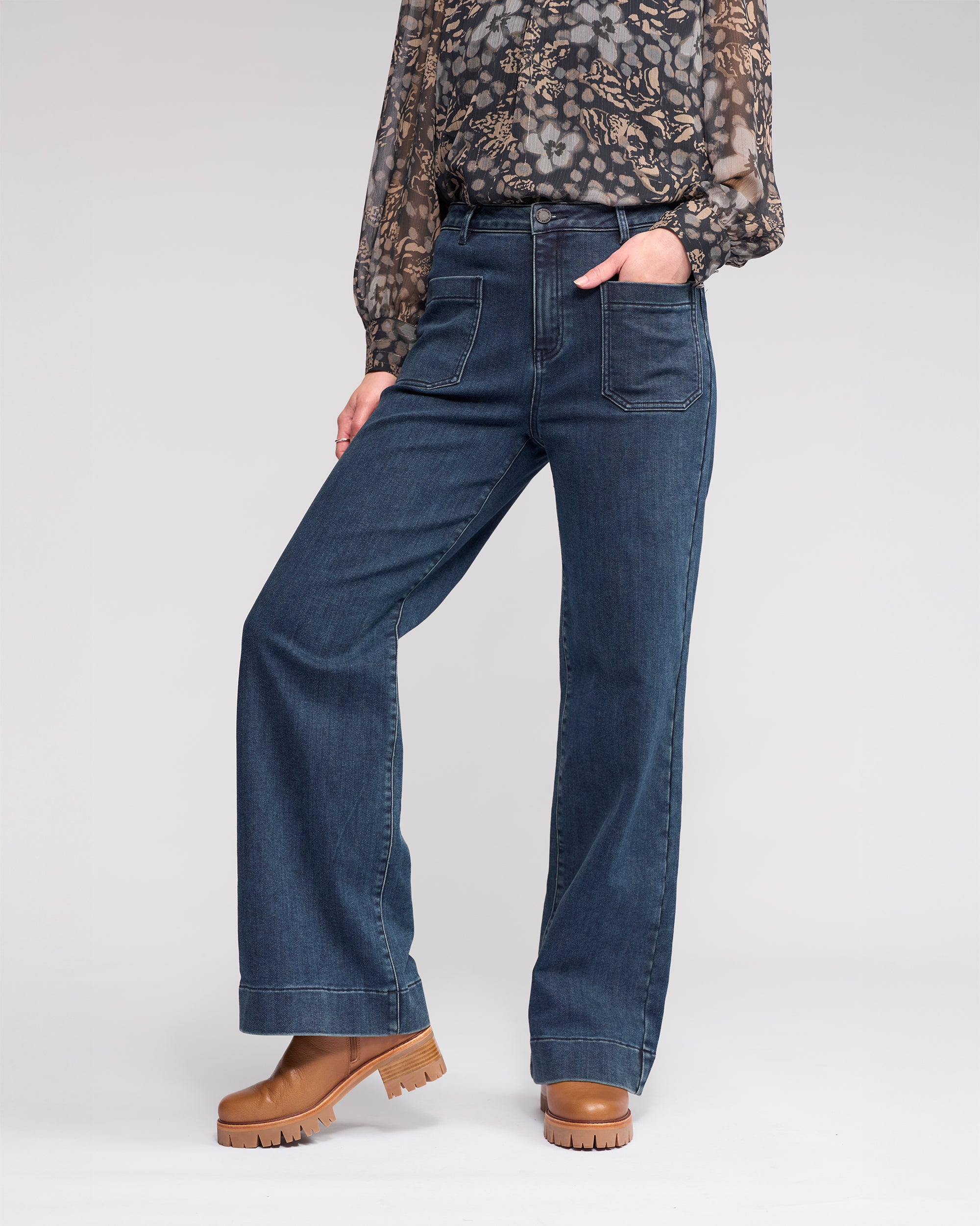A person wears the Knewe Label Roxie Jean in Slate Denim, featuring wide legs and patch pockets, styled with a sheer floral blouse and tan platform boots, standing with one hand in their pocket against a plain background.