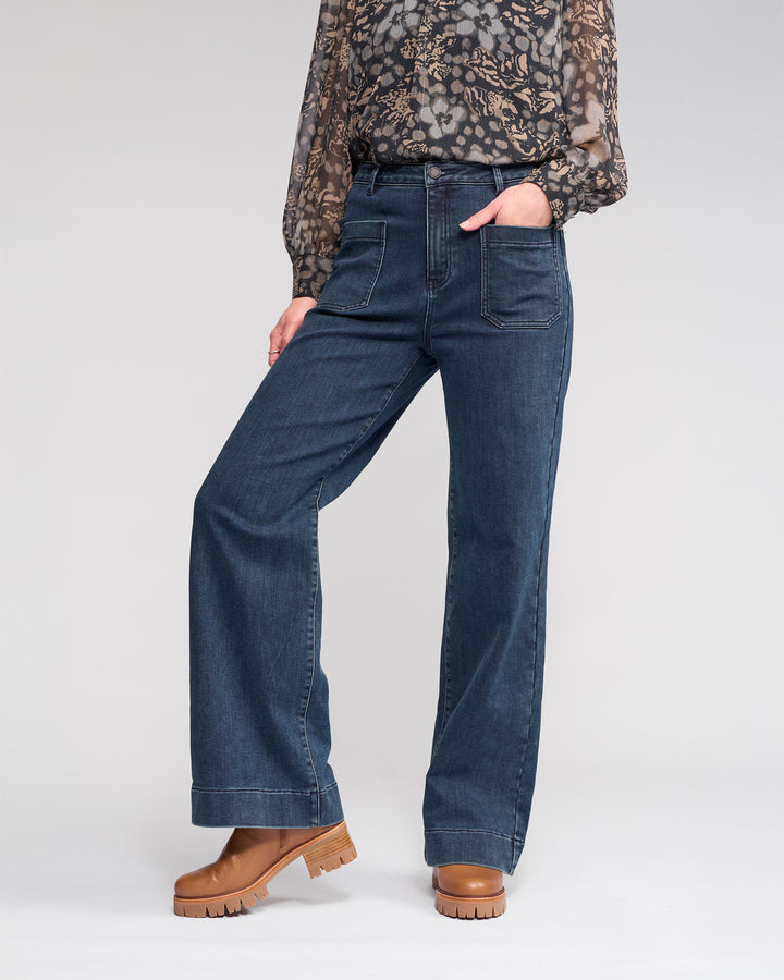 A person wears the Knewe Label Roxie Jean in Slate Denim, featuring wide legs and patch pockets, styled with a sheer floral blouse and tan platform boots, standing with one hand in their pocket against a plain background.