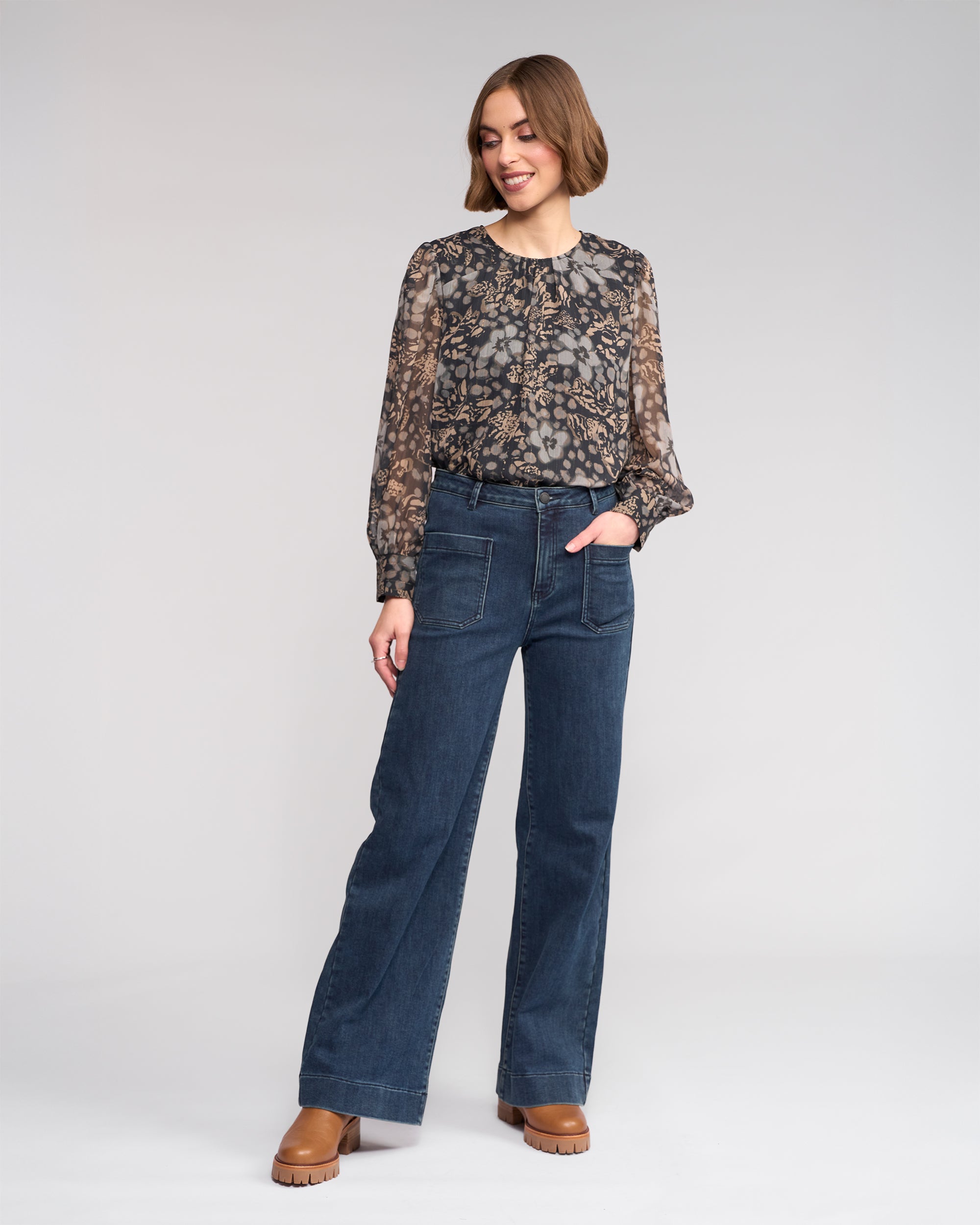 A woman with short hair smiles to the side, modeling Knewe Label's Roxie Jean in slate denim—wide-leg jeans with patch pockets—paired with a sheer floral blouse and brown platform shoes against a plain background.
