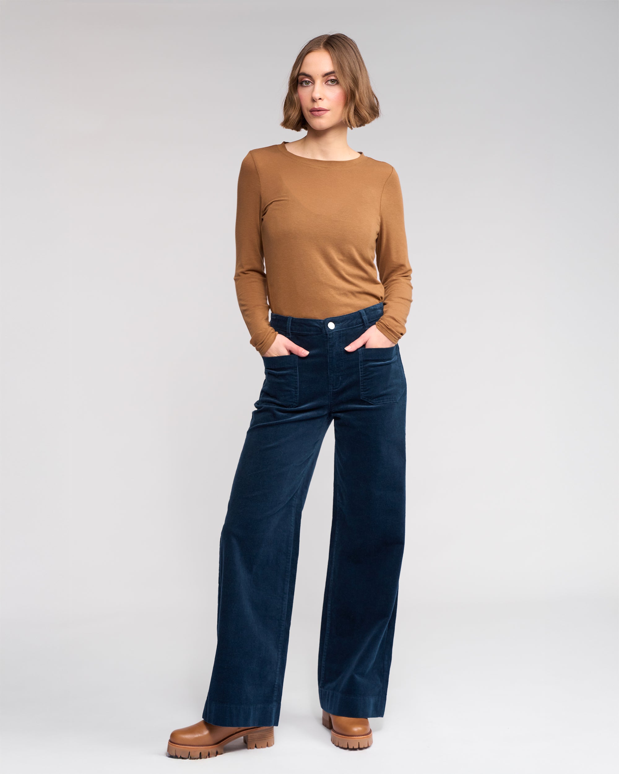 A woman stands in a neutral pose with hands in her pockets, wearing Knewe Label’s Roxie Cord - Airforce wide-leg pants, a brown long-sleeve top, and brown platform shoes against a light gray background.