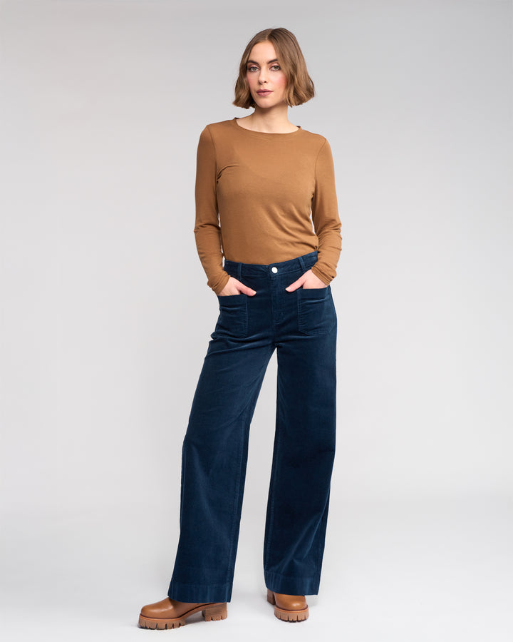 A woman stands in a neutral pose with hands in her pockets, wearing Knewe Label’s Roxie Cord - Airforce wide-leg pants, a brown long-sleeve top, and brown platform shoes against a light gray background.