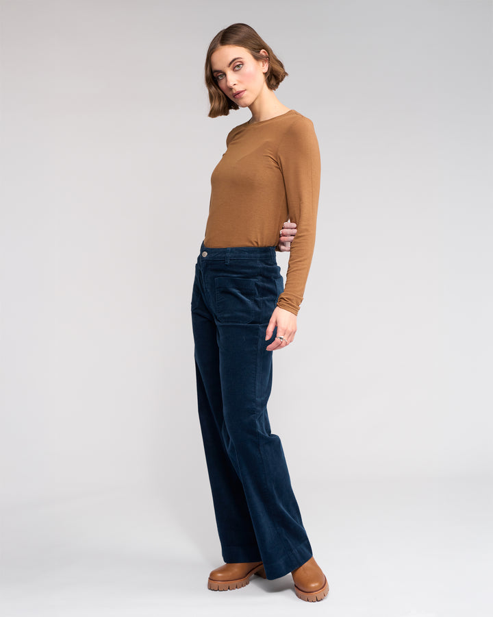 A person with short brown hair wears a fitted brown long-sleeve shirt, Knewe Label’s Roxie Cord - Airforce dark blue wide-leg corduroy pants, and brown platform shoes, standing against a plain light gray background.