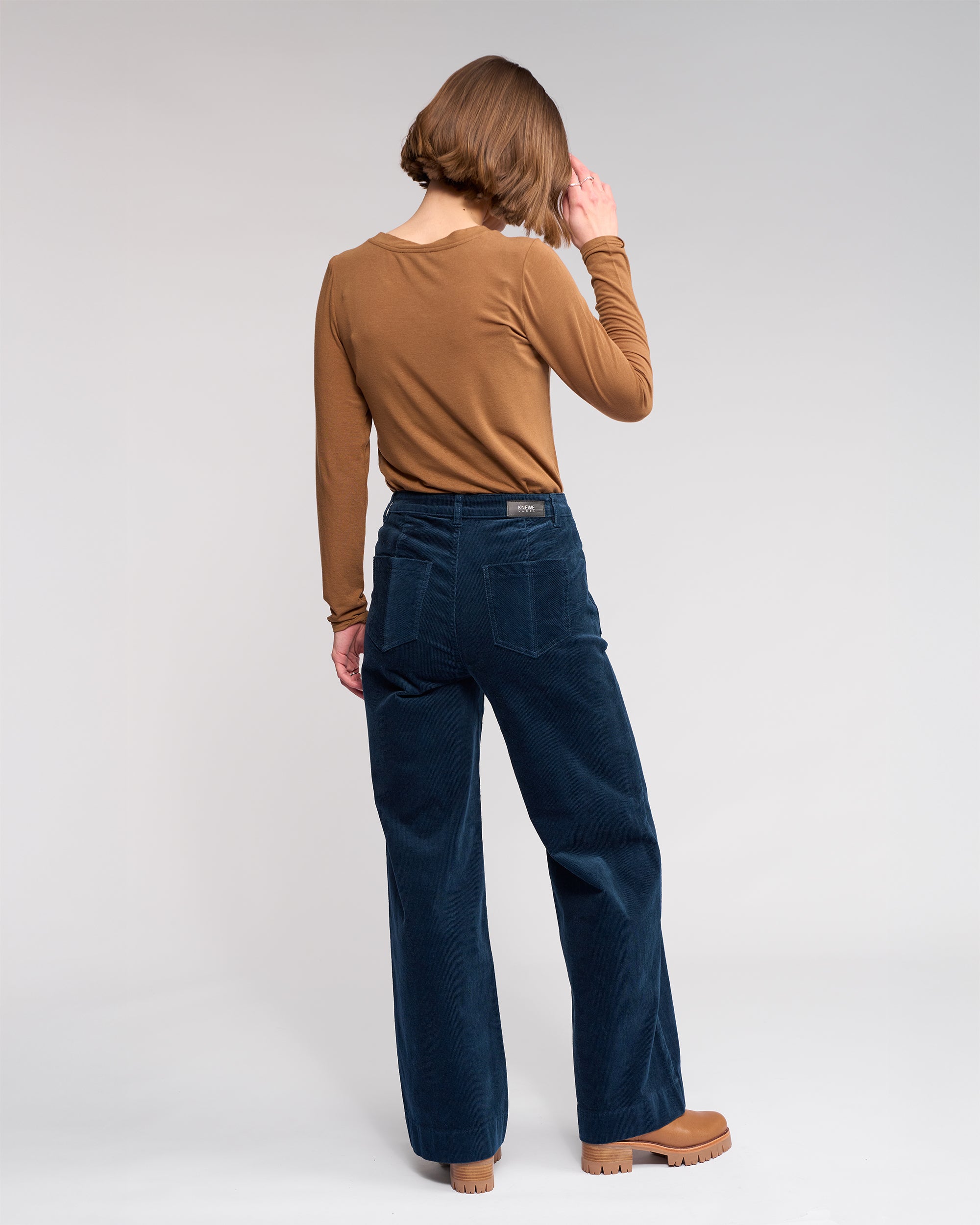 A person with short brown hair faces away, wearing a brown long-sleeve top, tan shoes, and Knewe Label Roxie Cord - Airforce dark blue wide-leg corduroy pants against a plain light background.
