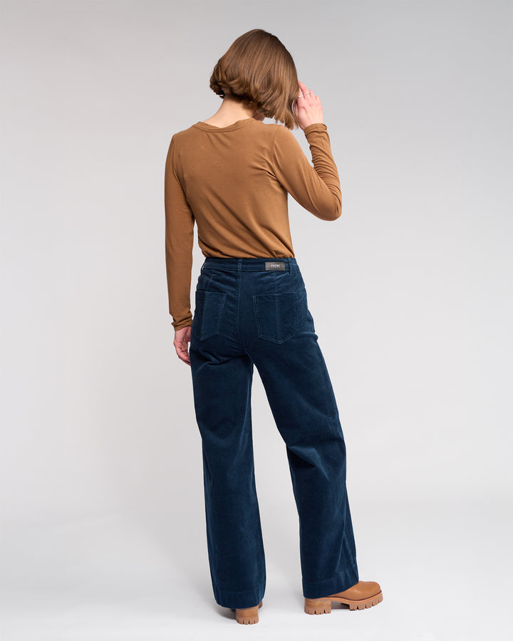 A person with short brown hair faces away, wearing a brown long-sleeve top, tan shoes, and Knewe Label Roxie Cord - Airforce dark blue wide-leg corduroy pants against a plain light background.