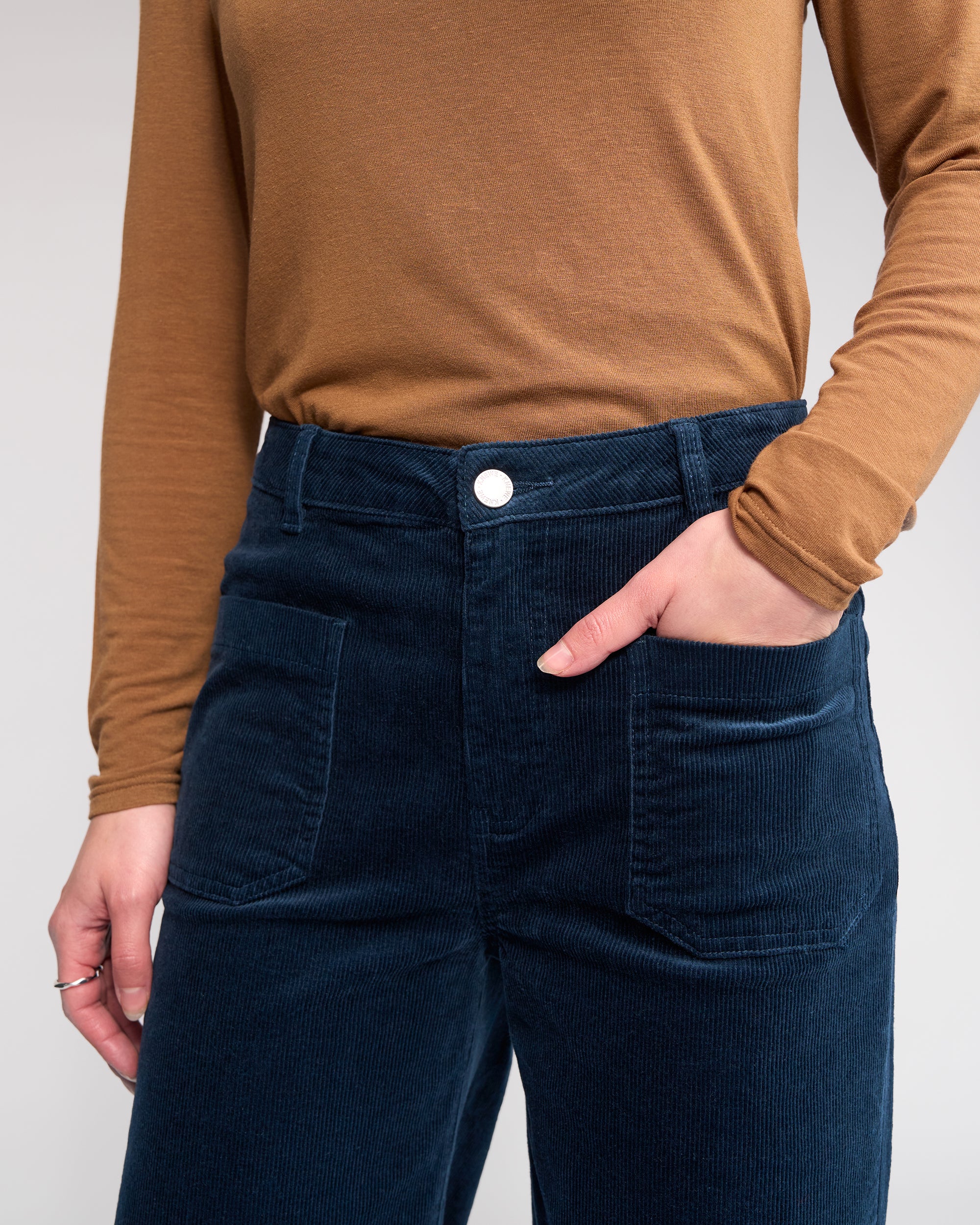 A person wears the Knewe Label Roxie Cord - Airforce wide-leg pants, made of wide wale cord in dark blue, paired with a tan long-sleeve shirt, standing with one hand in their pocket against a neutral background.