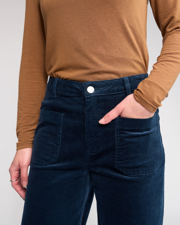 A person wears the Knewe Label Roxie Cord - Airforce wide-leg pants, made of wide wale cord in dark blue, paired with a tan long-sleeve shirt, standing with one hand in their pocket against a neutral background.