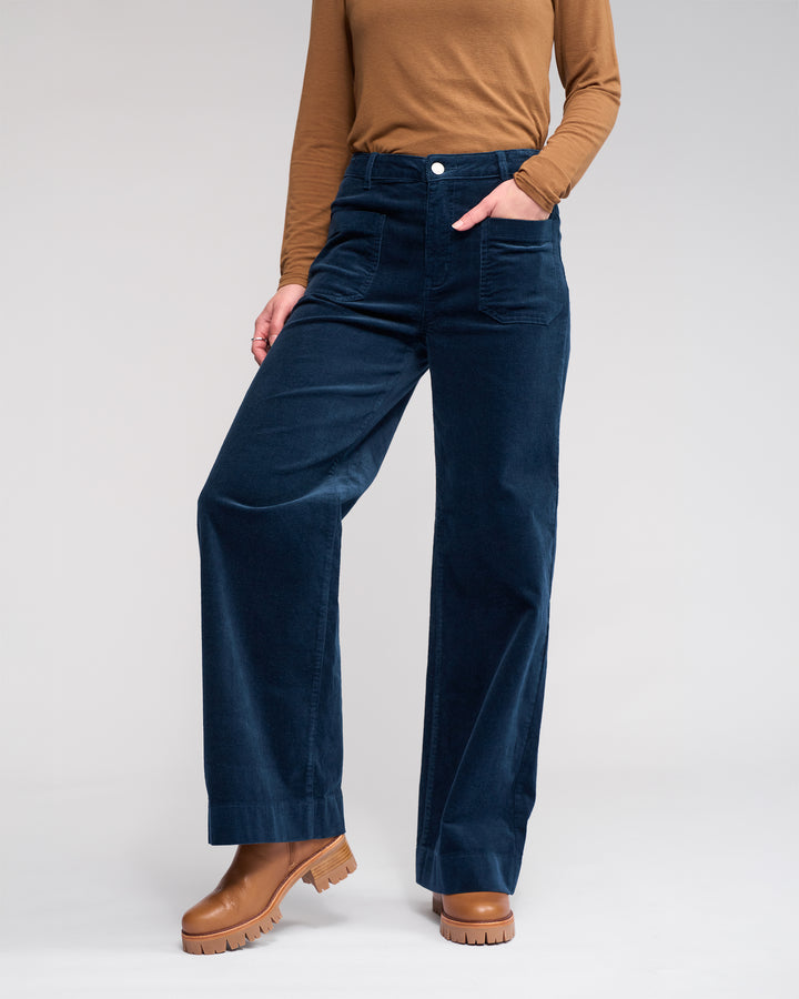 A person stands against a plain background, wearing a brown long-sleeve top, brown ankle boots, and the Knewe Label Roxie Cord - Airforce wide-leg pants in dark blue with one hand in their pocket.