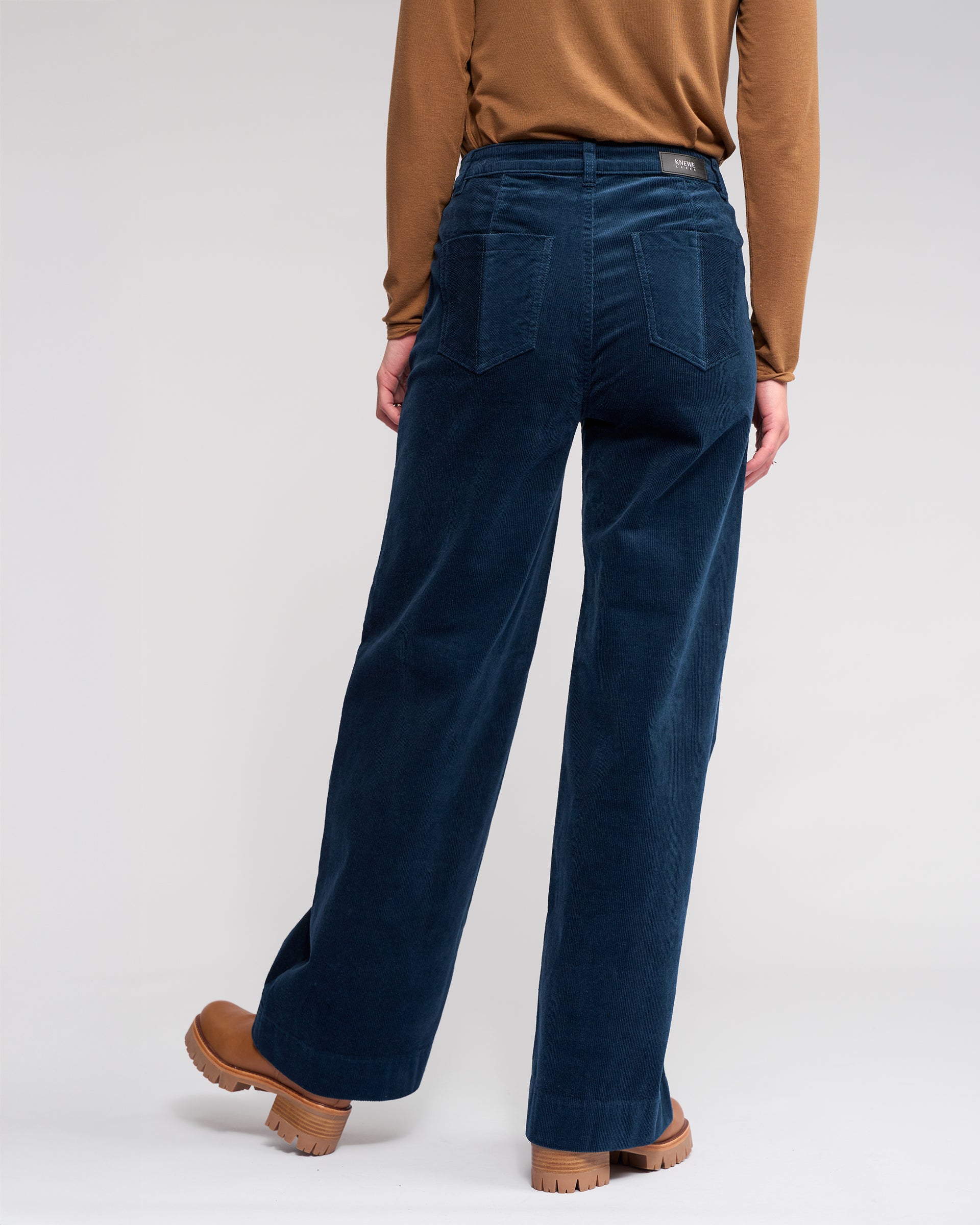 A person in a brown long-sleeve shirt, Knewe Label Roxie Cord - Airforce wide-leg pants, and tan platform shoes stands with their back to the camera against a light gray background.