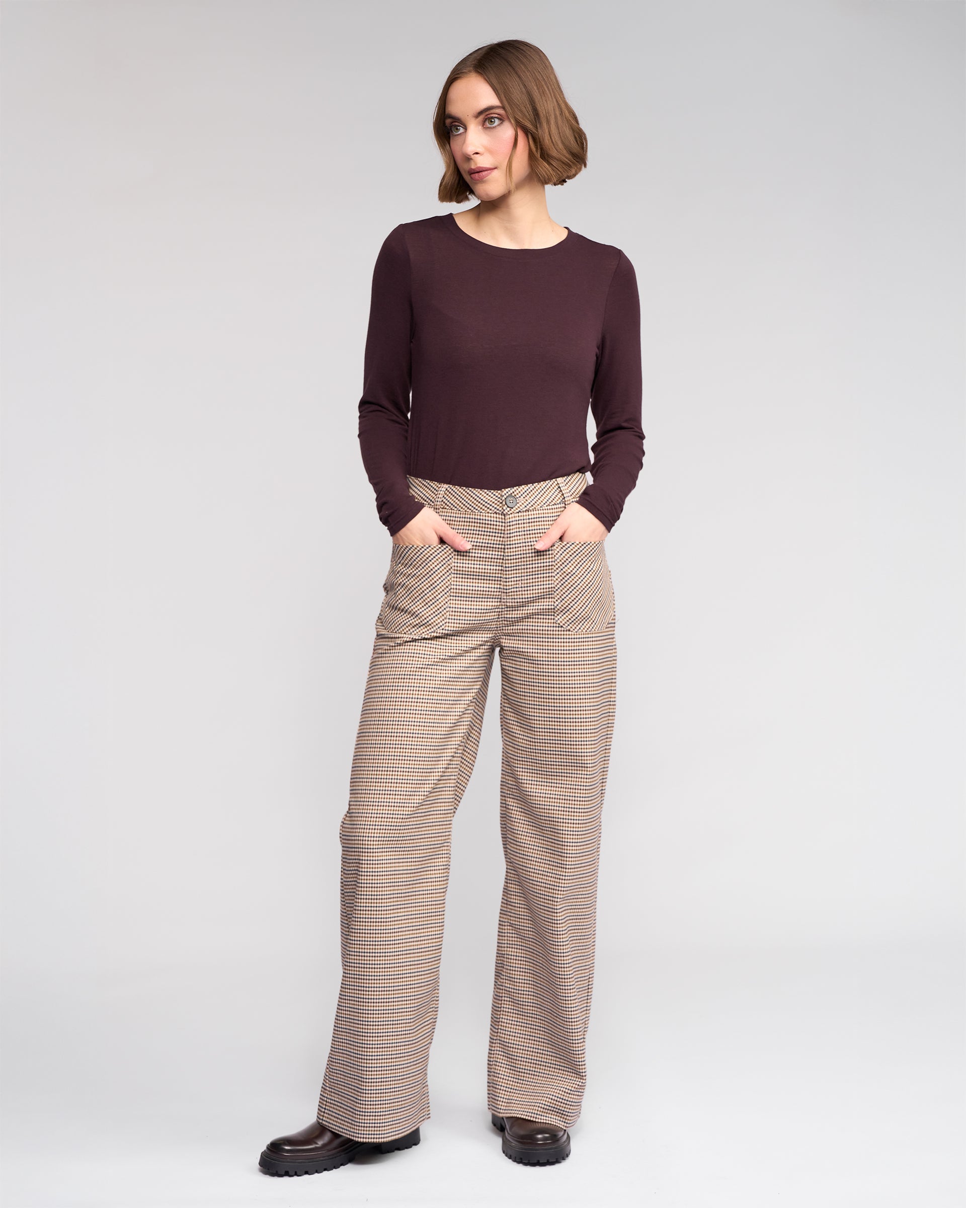 A woman stands against a plain background, wearing the Roxie Pant by Knewe Label—a high-waisted, wide-leg checked style—with her hands in her pockets and looking slightly to the side.