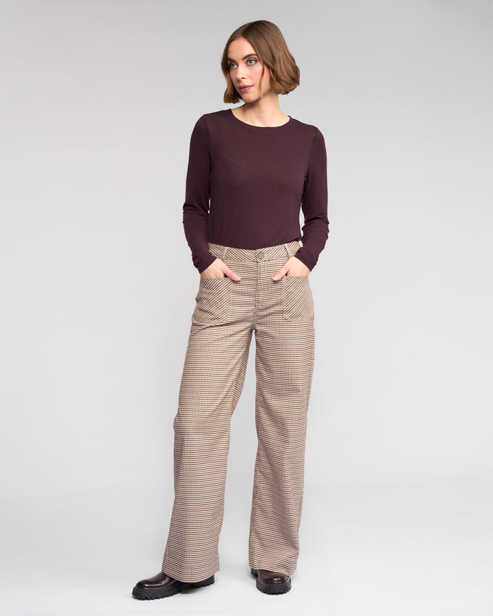 A woman stands against a plain background, wearing the Roxie Pant by Knewe Label—a high-waisted, wide-leg checked style—with her hands in her pockets and looking slightly to the side.